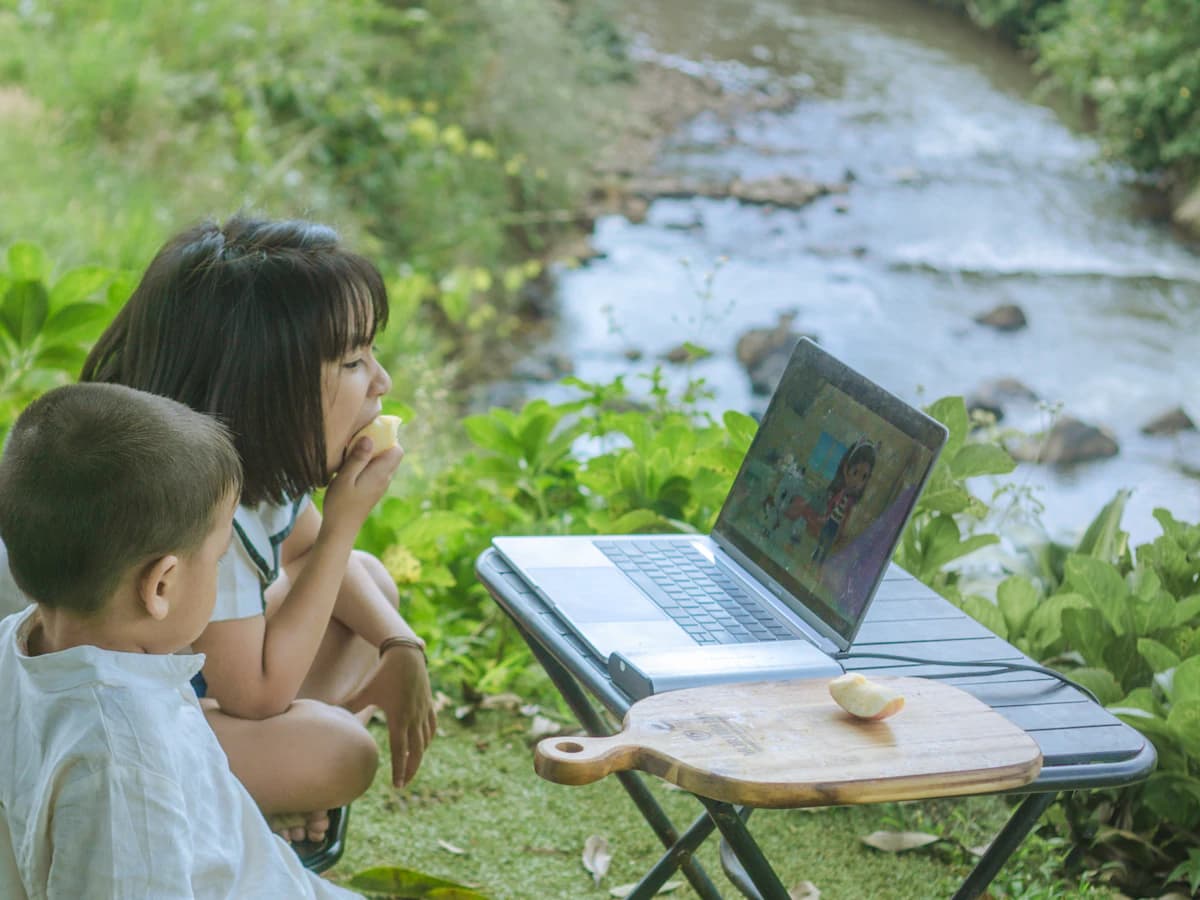 Two people sharing an outdoor table with a laptop by a stream